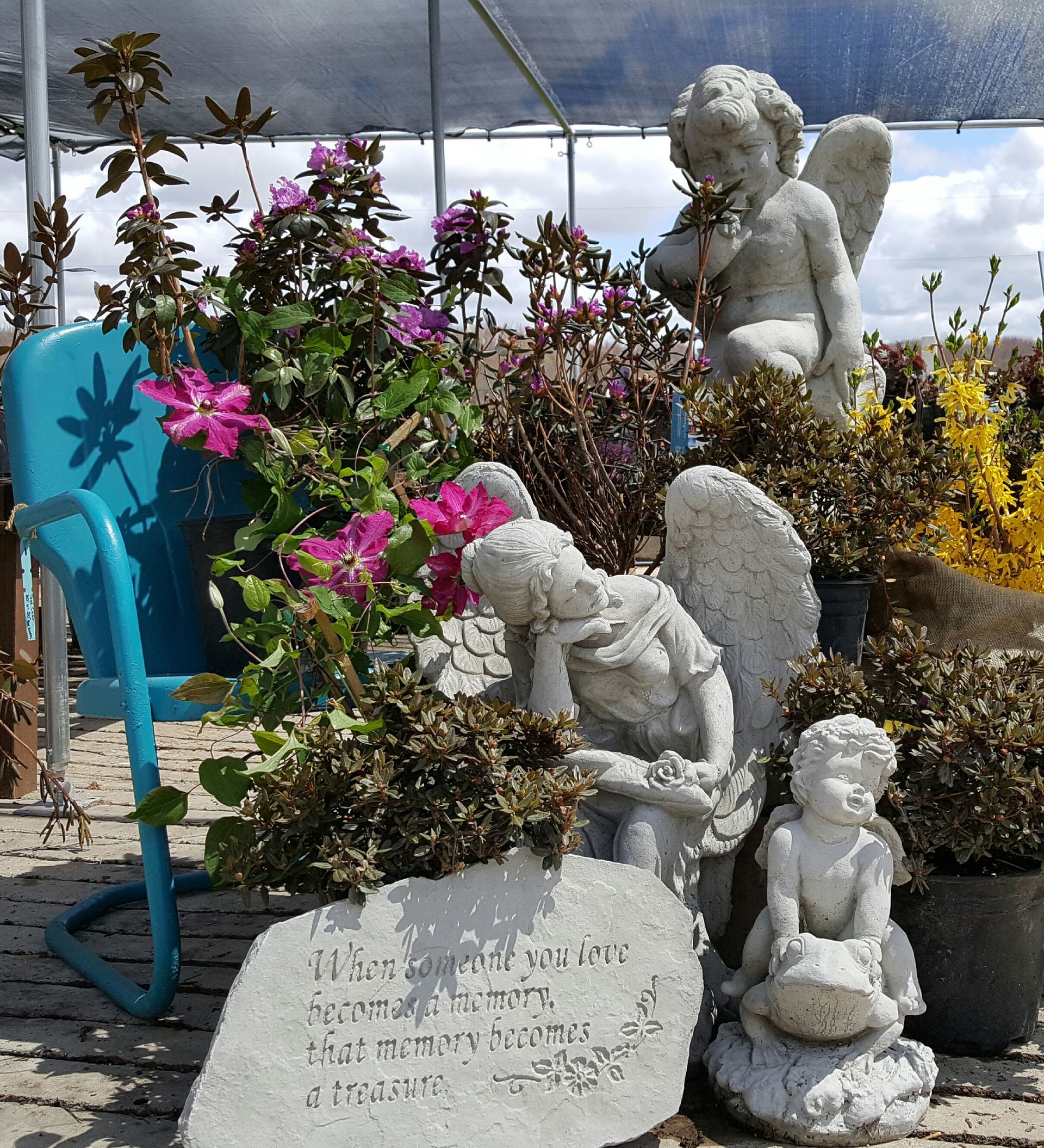 Three angel statues amongst flowers and foliage, with a blue chair, outdoors.