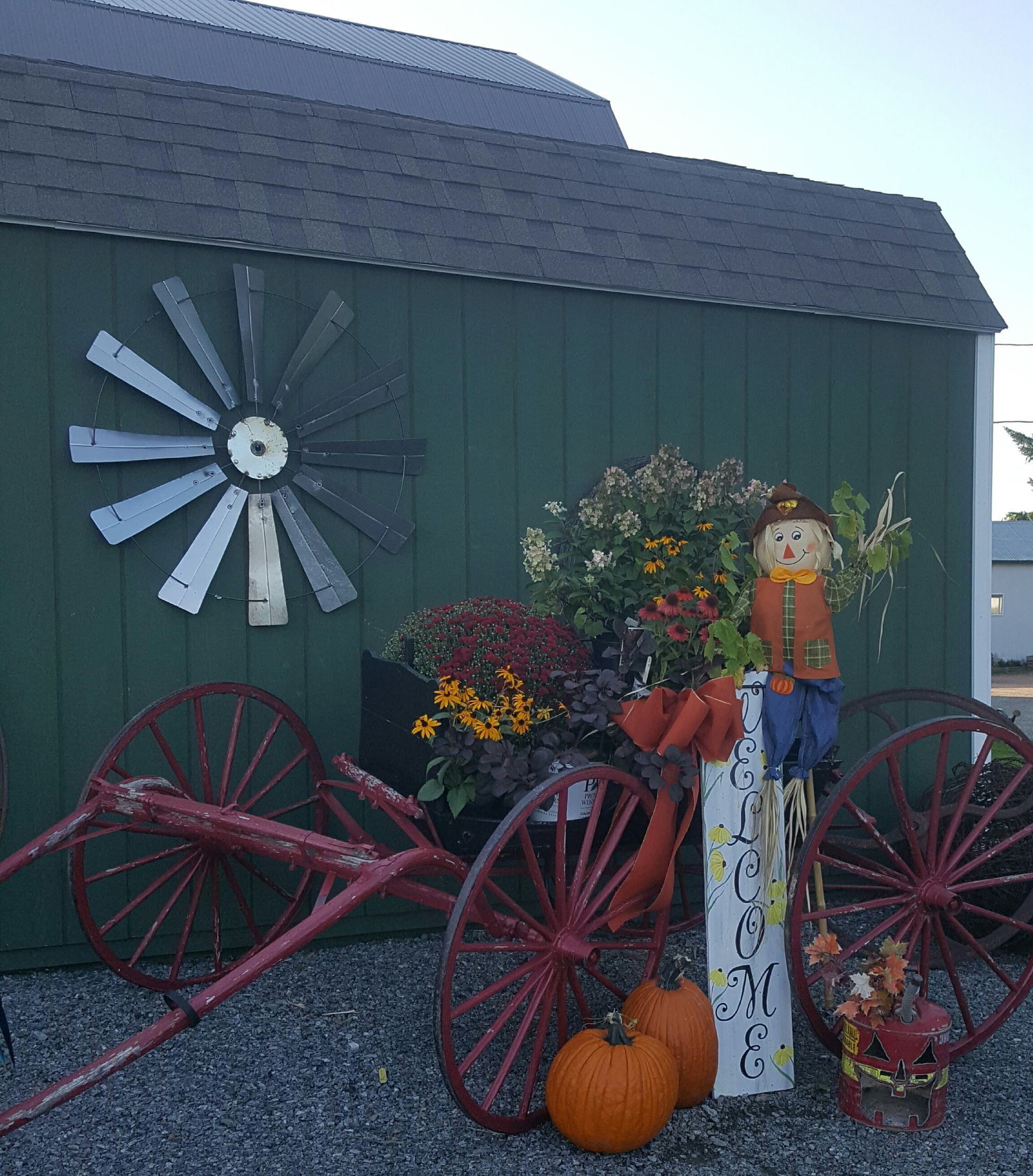 Fall-themed display with pumpkins, scarecrow, wagon wheels, and a windmill decoration against a green building.