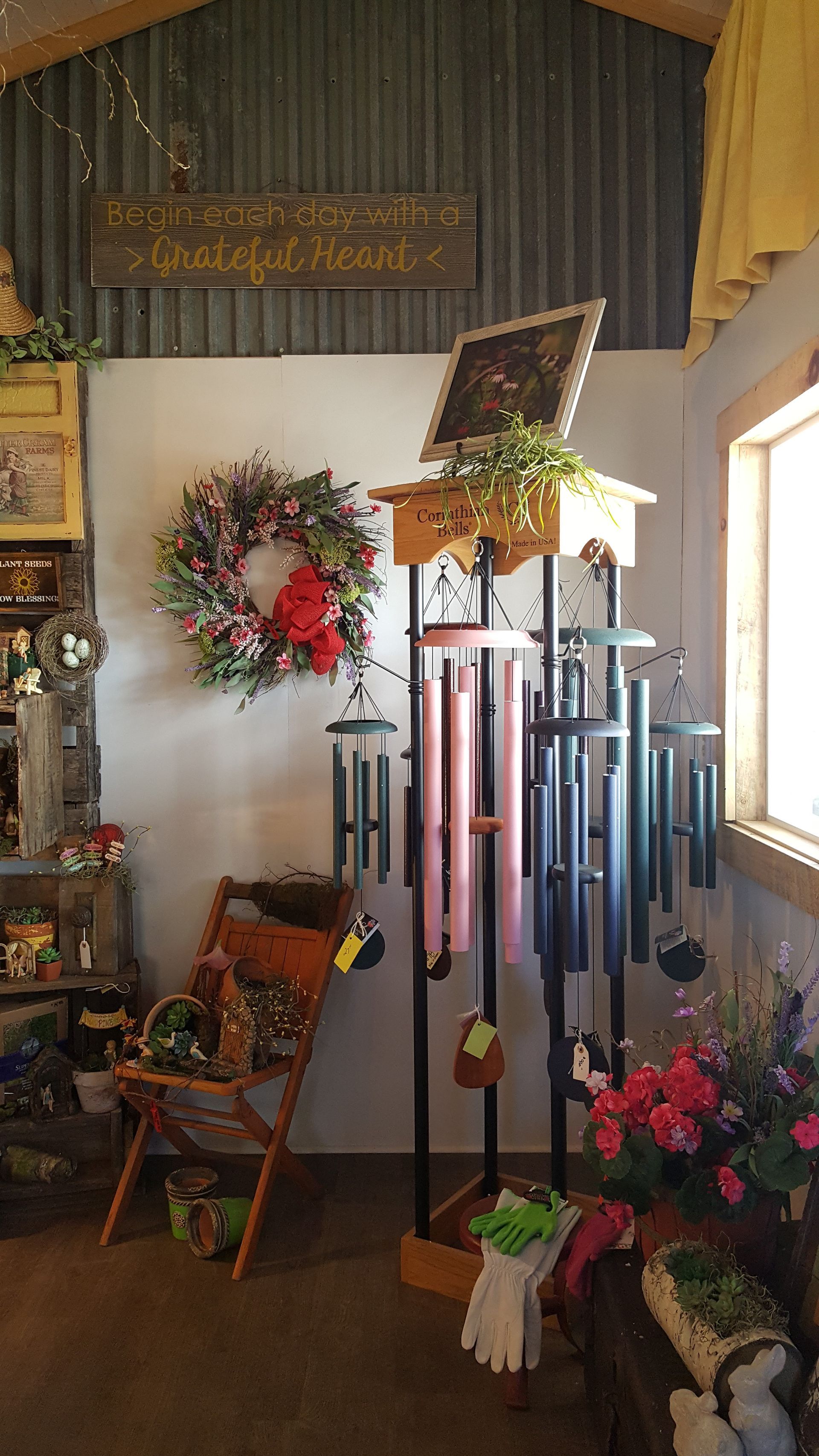 Indoor display of gifts: wind chimes, wreath, chair, plants. Metal shed interior with white wall.