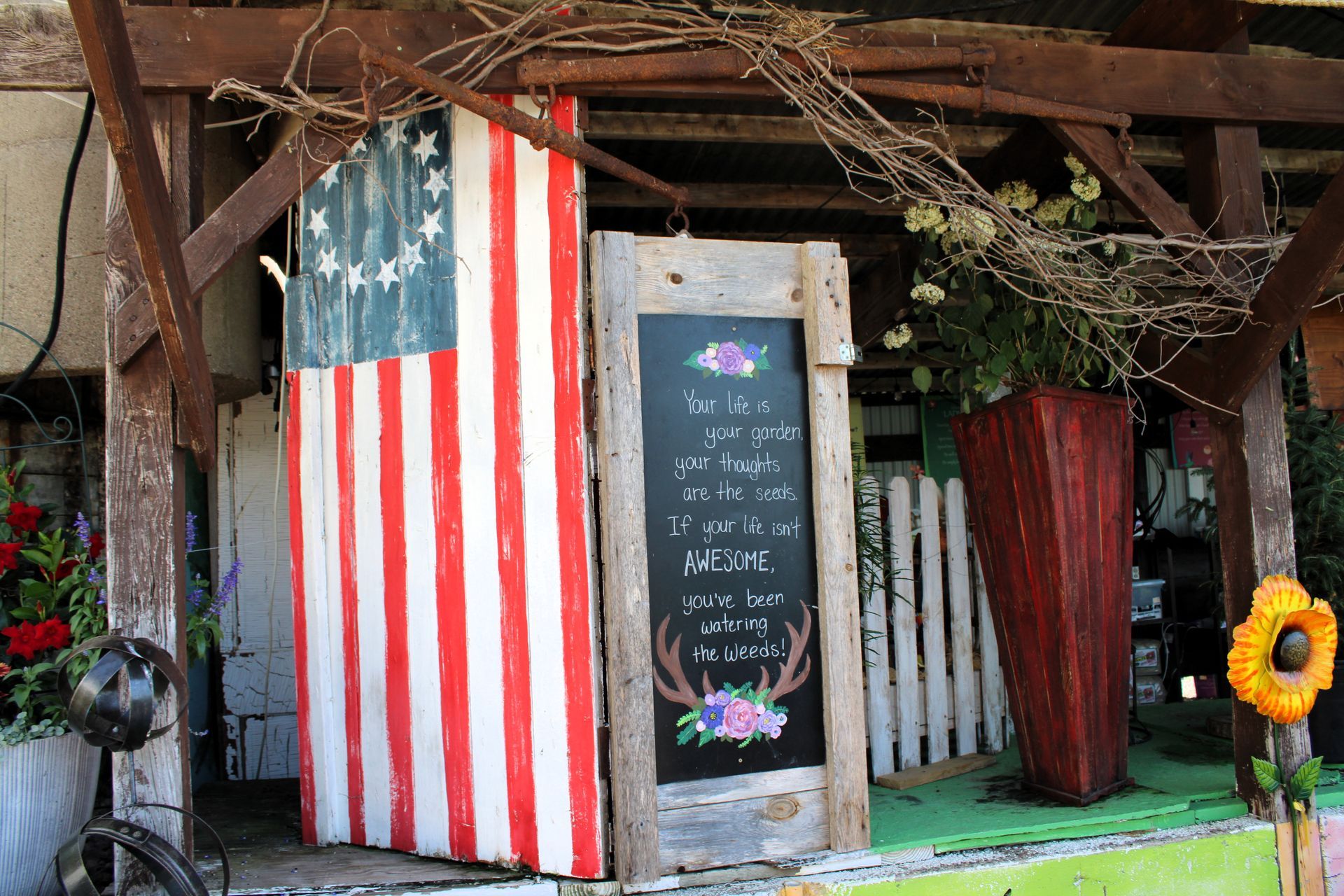 Hand-painted American flag, door with text, wooden structure, flowers, and rustic decor.
