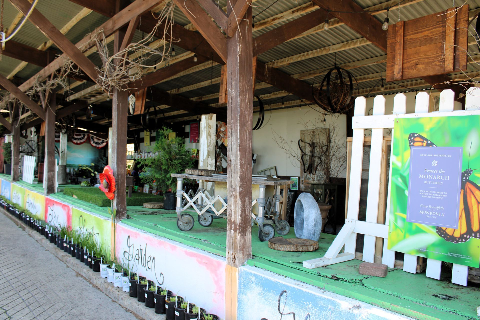 Covered outdoor display of plants, art, and signs; under wooden beams and corrugated metal roof.