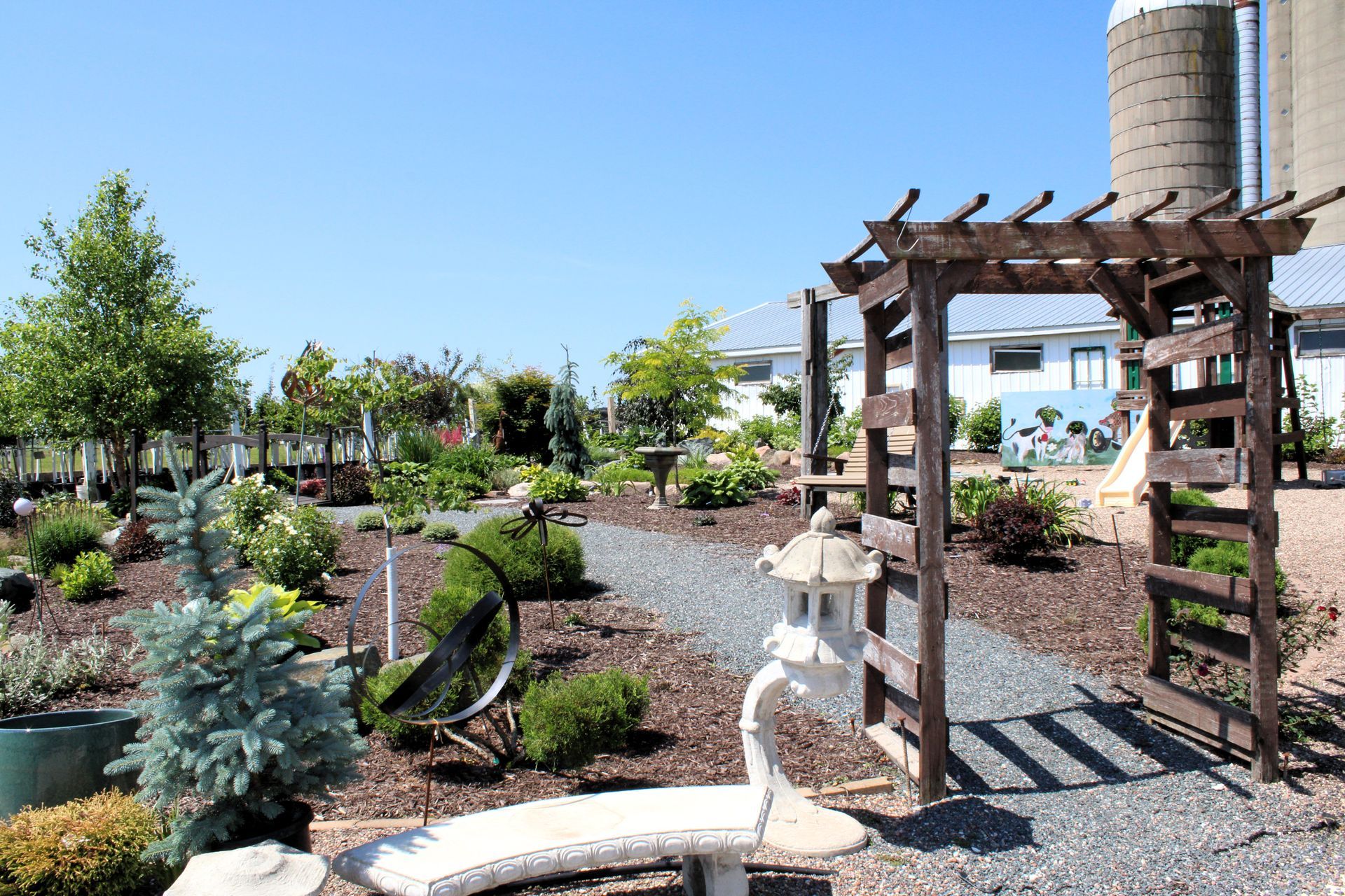Garden scene with a wooden archway, stone lantern, and gravel pathway leading towards plants and a silo.