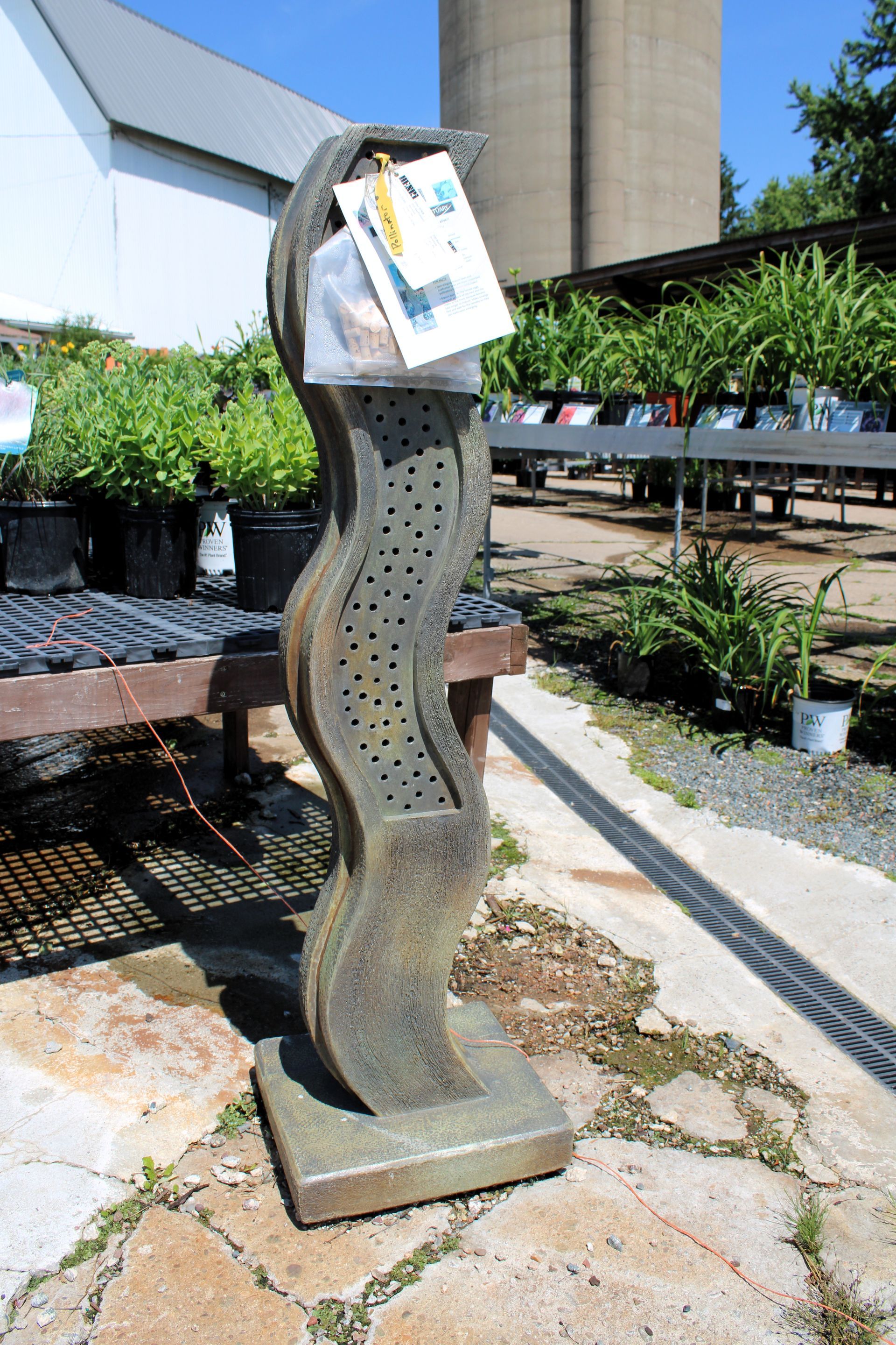 Abstract, wavy sculpture with small holes holds papers outdoors near plants and a storage building.