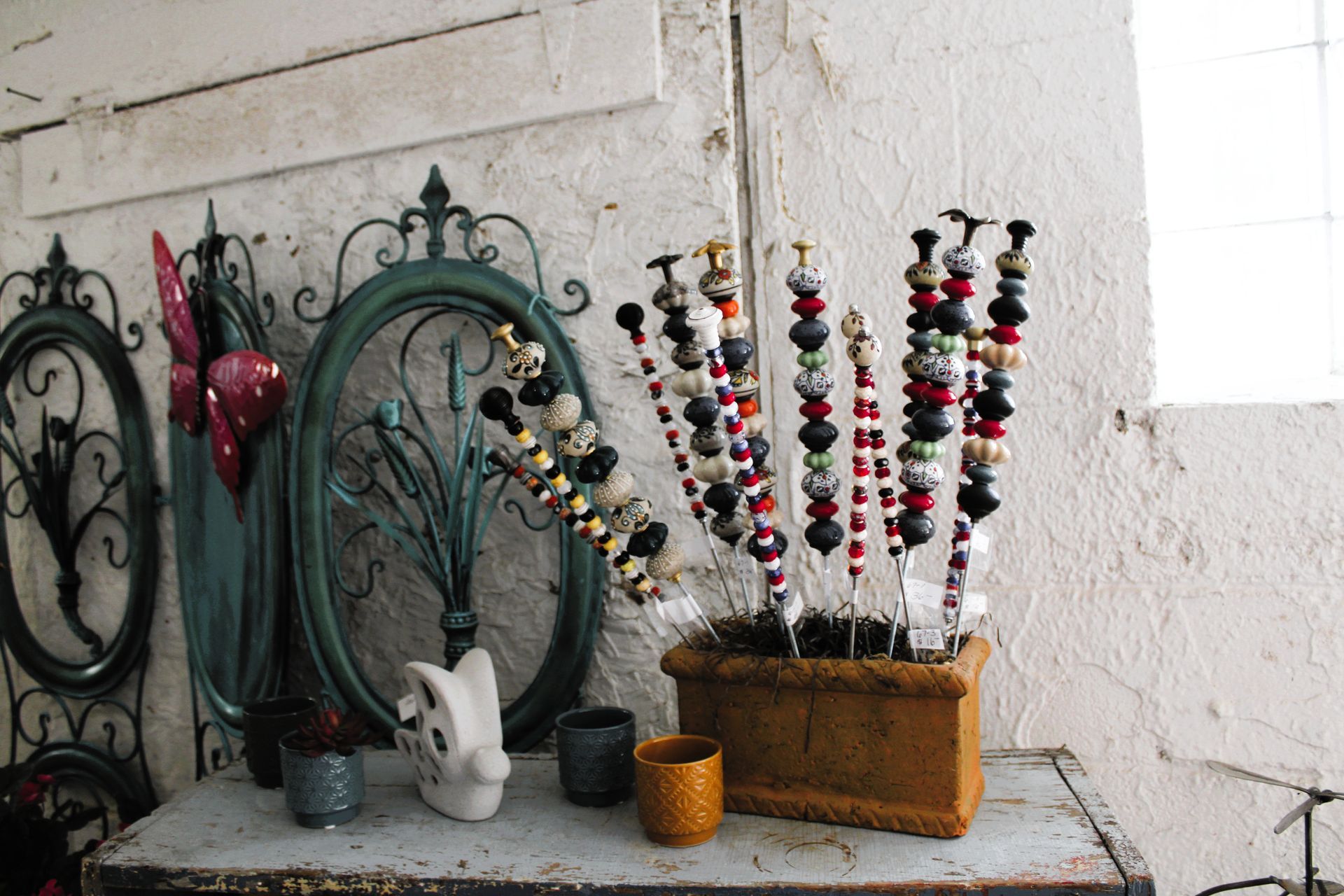 Display of decorative beaded stems in a planter on a weathered metal table, with ornate metal frames against a textured wall.