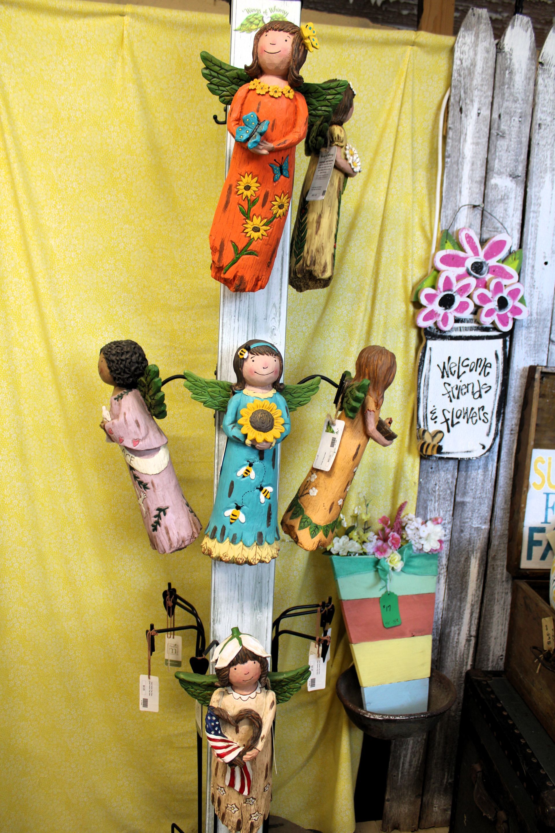 Angels hanging on a white post. Various colors, holding flowers.  Bucket of flowers below. Yellow backdrop.