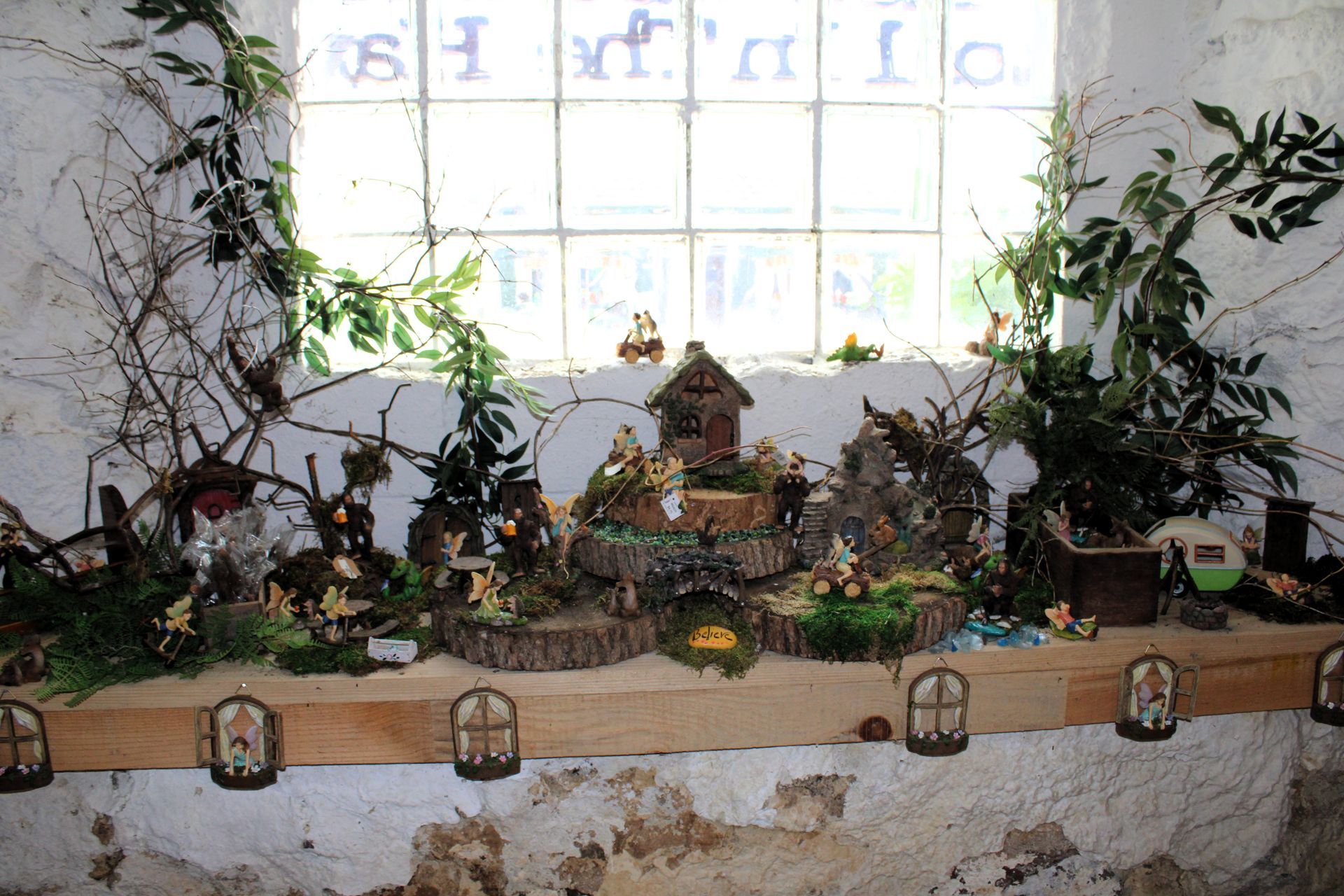 A whimsical fairy garden display on a wooden ledge beneath a window. Features tiny houses, figurines, and greenery.