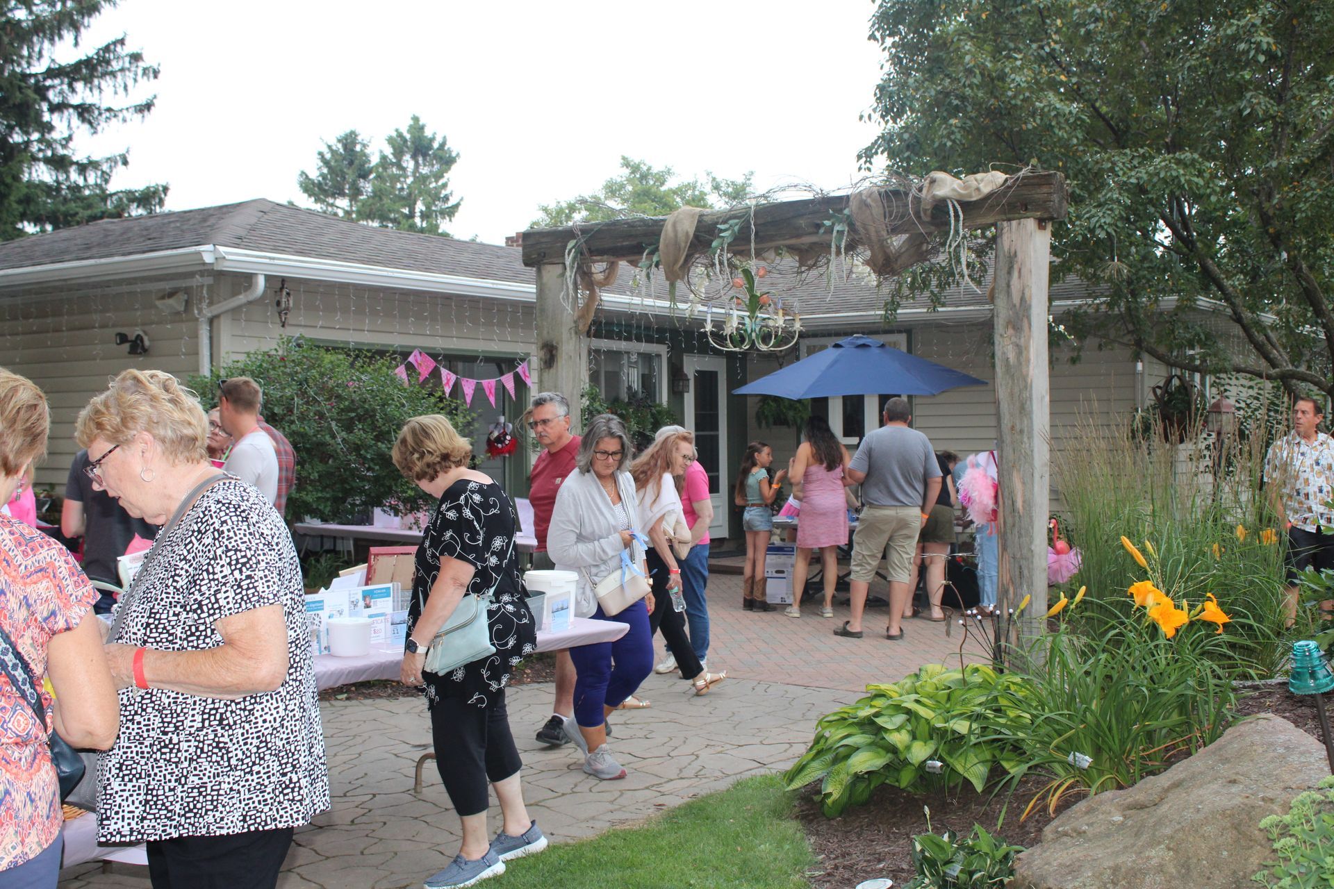 People gather outdoors at a party, near a building with an arbor and a table, some are eating or socializing.