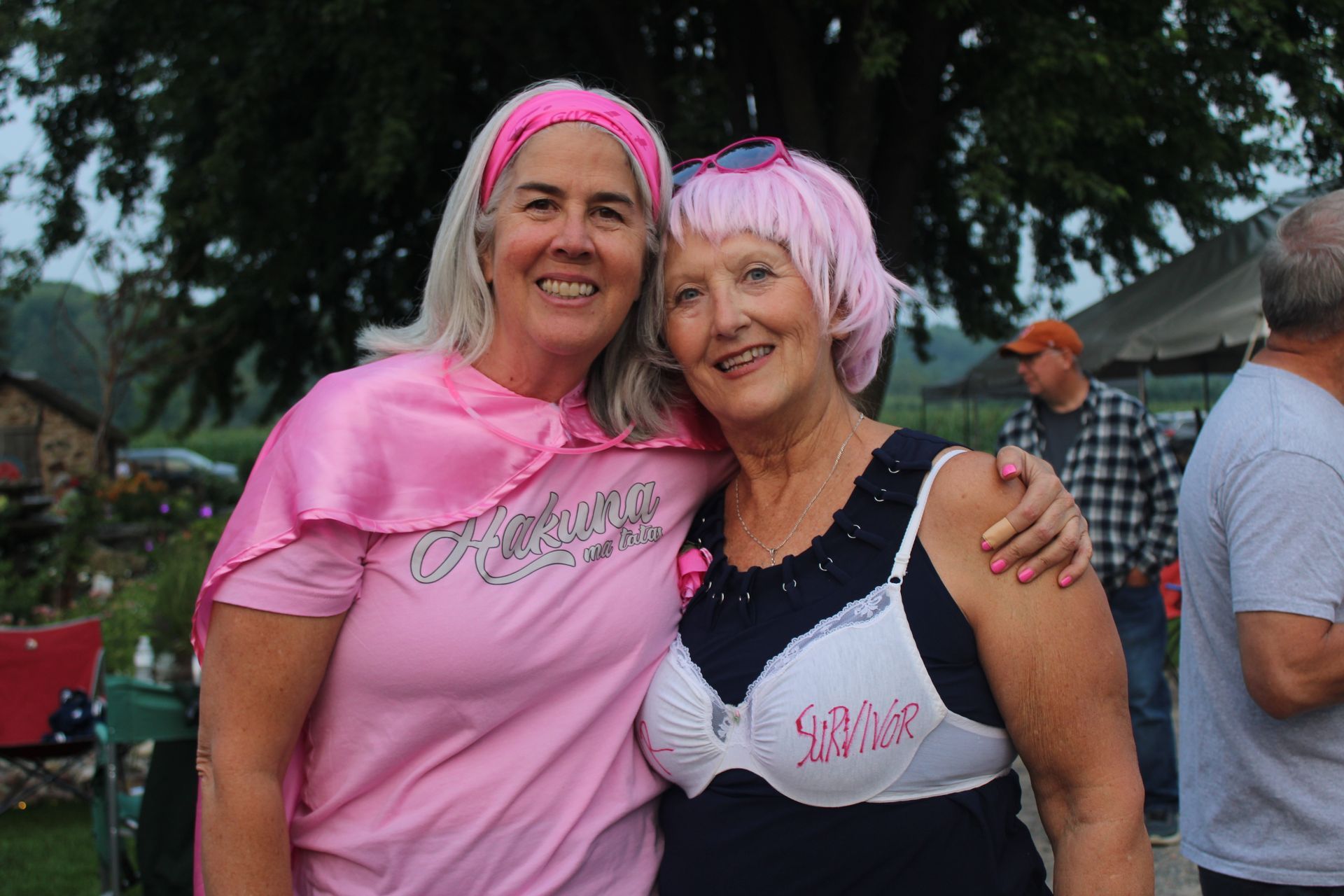 Two women in pink, one wearing a bra, smiling. Outdoors, trees, other people in background.