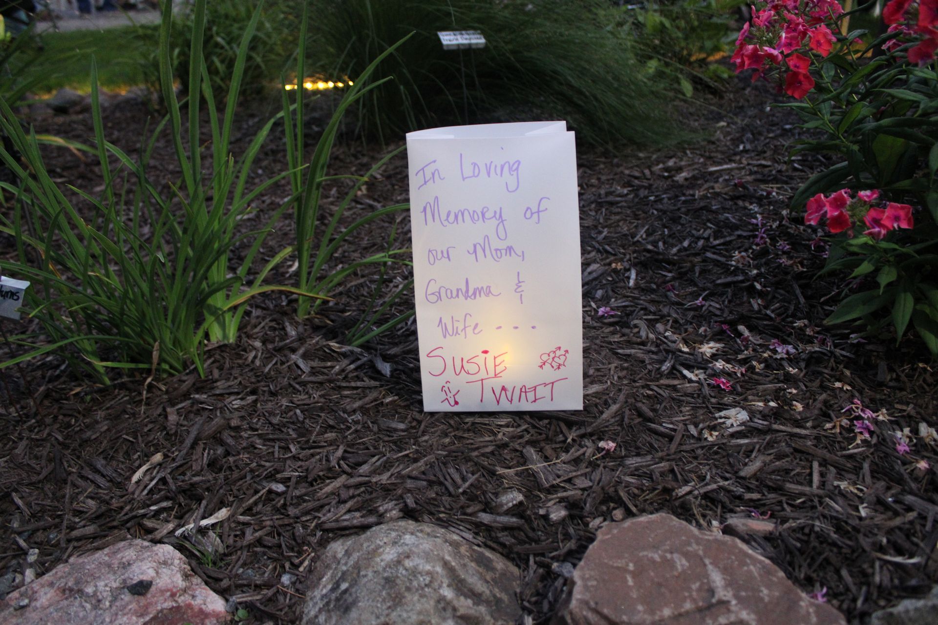 A luminaria with handwritten text honoring someone, placed in a garden bed with rocks, mulch, and greenery.