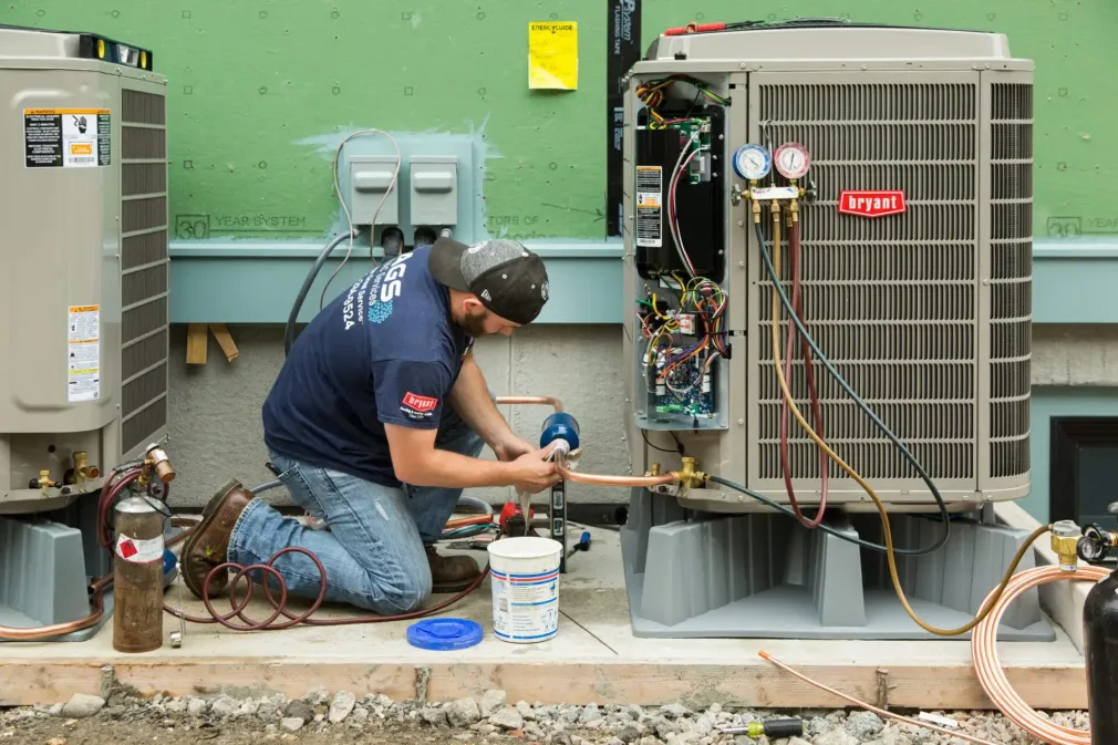 Technician kneels beside outdoor HVAC units, repairing wiring with tools and a bucket nearby.