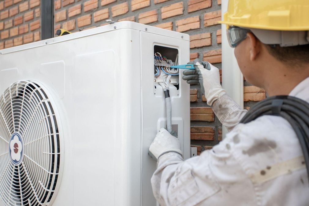 Technician wiring a white outdoor air conditioner unit beside a brick wall