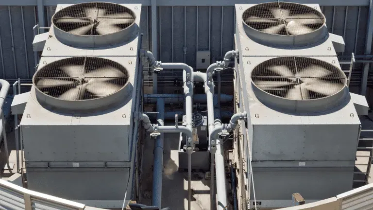 Four rooftop HVAC units with large fans connected by pipes on a gray industrial roof.