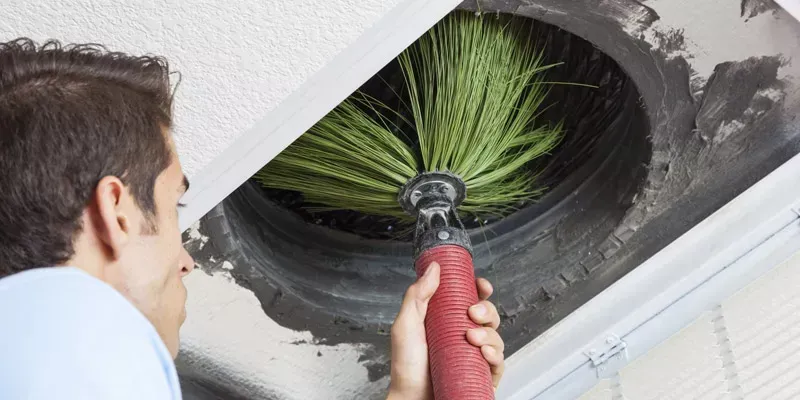 Man cleaning a ceiling vent with a vacuum brush, removing dust and debris