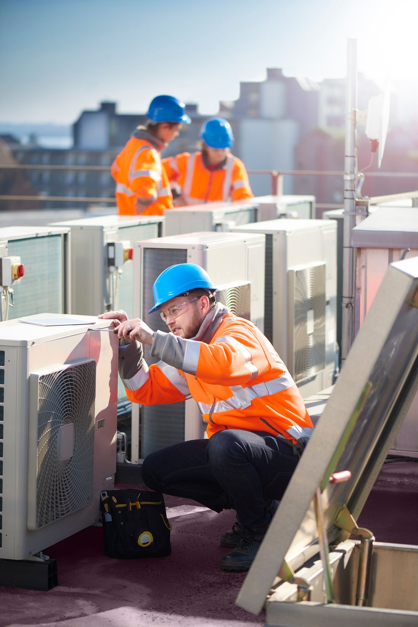 HVAC technician in orange safety vest and hard hat working on rooftop unit, two colleagues in background.