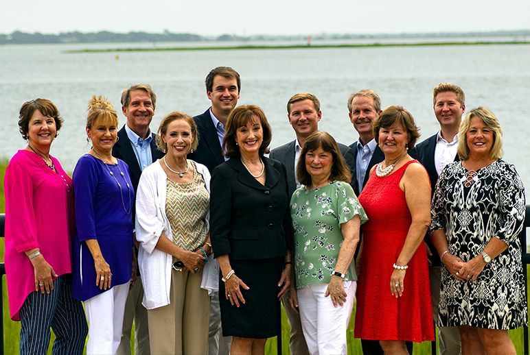 A group of twelve people stand in two rows smiling before a body of water on a sunny day.