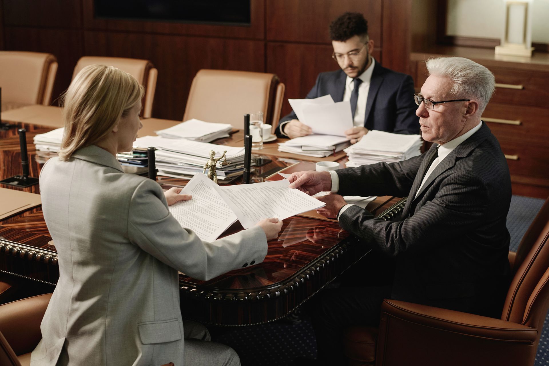 Two people sit at a desk reviewing documents in a formal office, with a third person observing in the background.