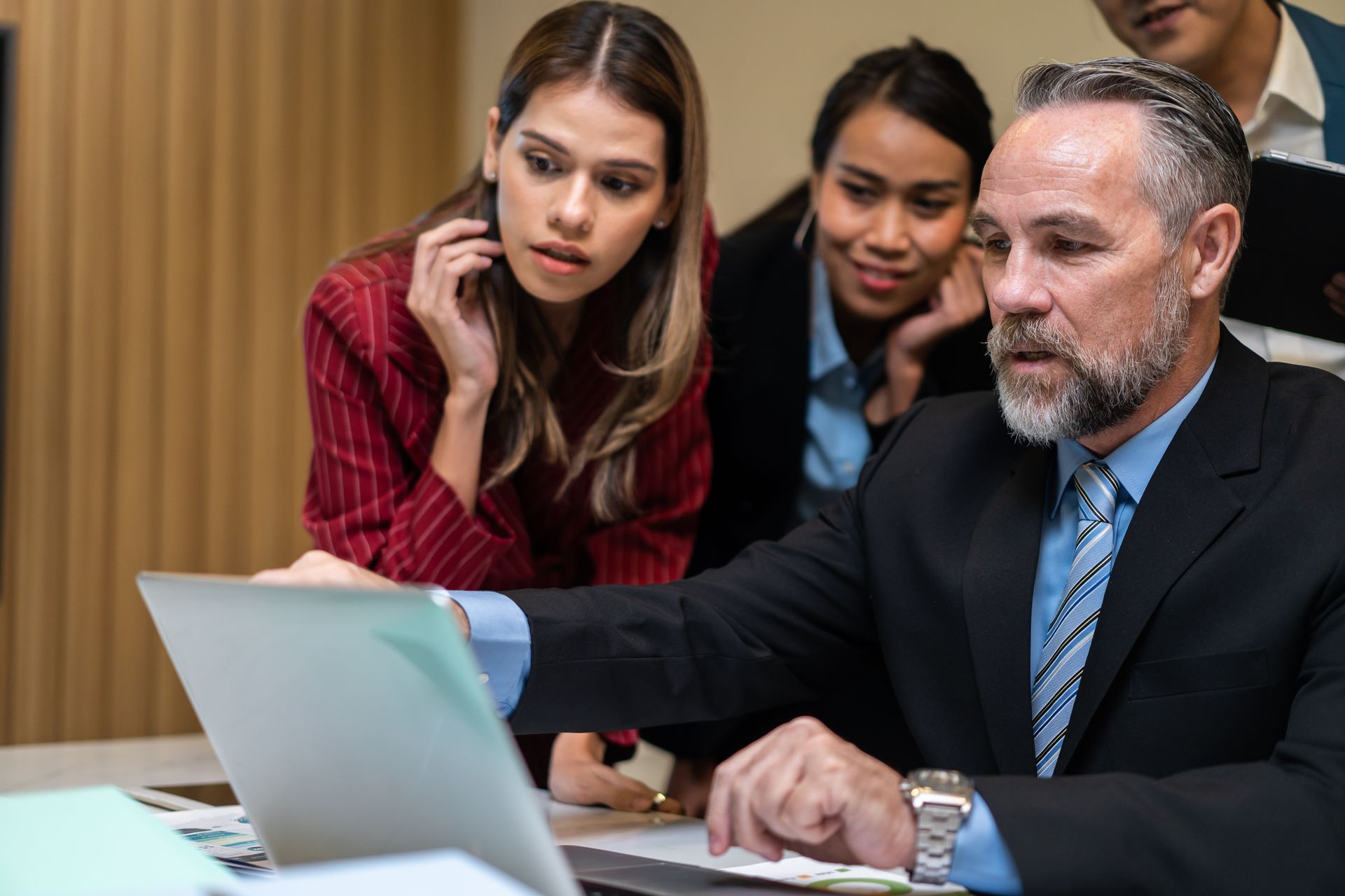 A professional team in business attire gathers around a laptop, focused on the screen during a collaborative discussion.