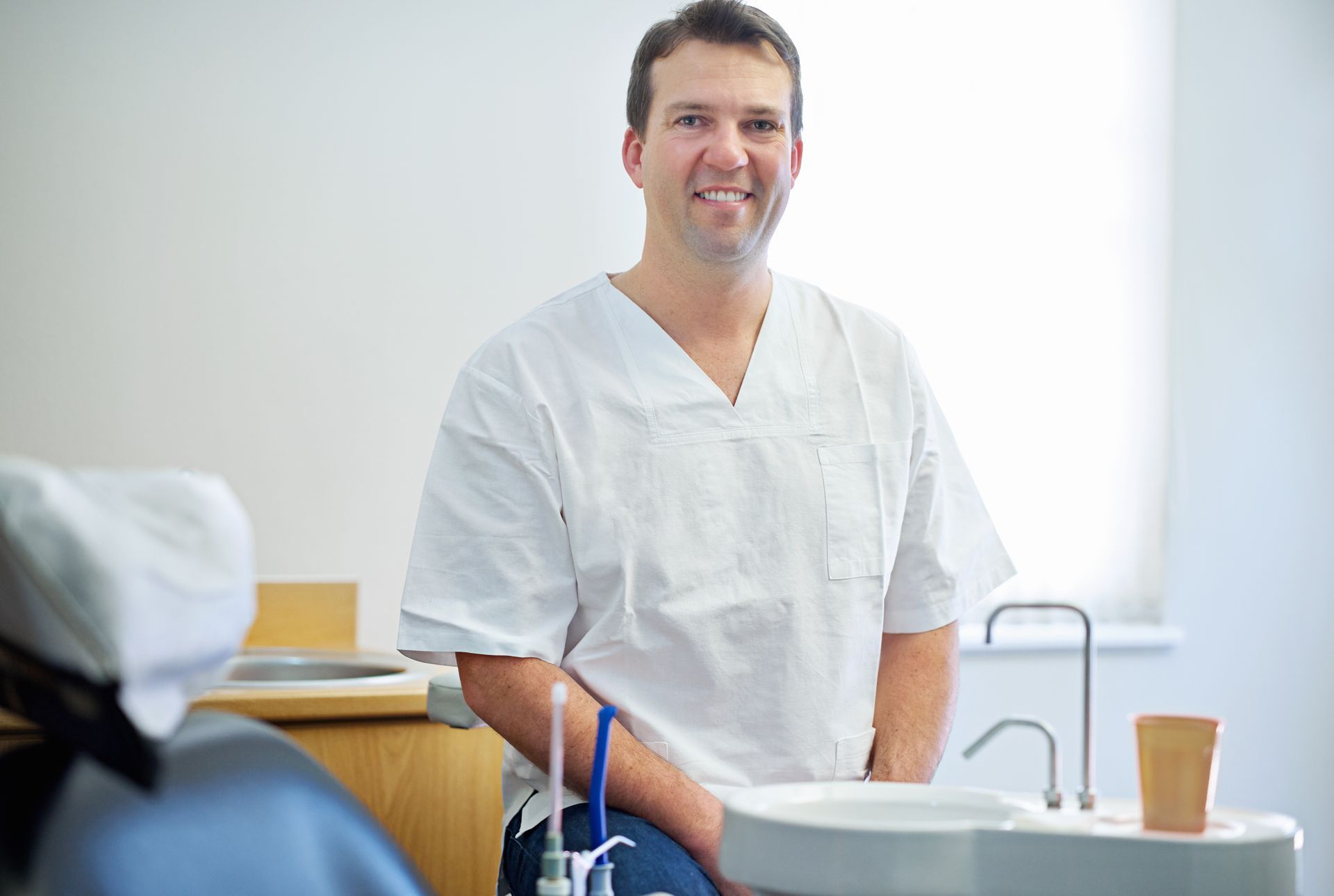 A smiling professional wearing white medical scrubs, sitting in a clean, brightly lit dental office.