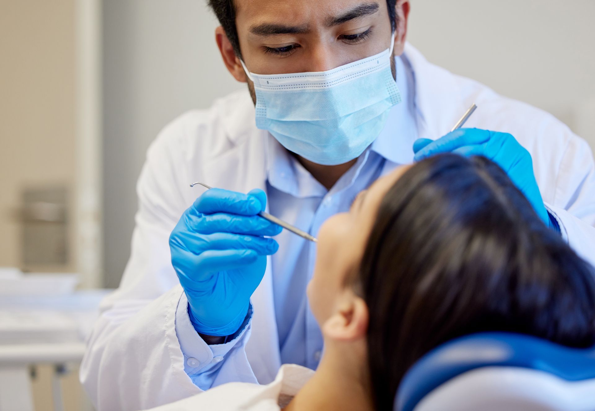 A dental professional wearing a mask and blue gloves examines a patient's teeth in a clinical setting.