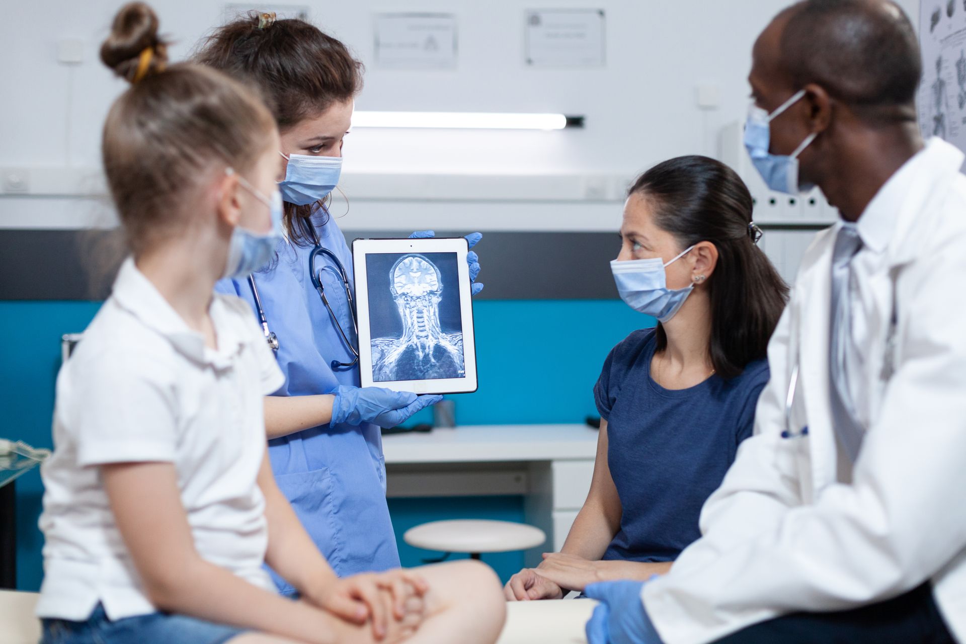 Medical staff in blue scrubs and white coats show a digital X-ray to a parent and child in a clinical examination room.
