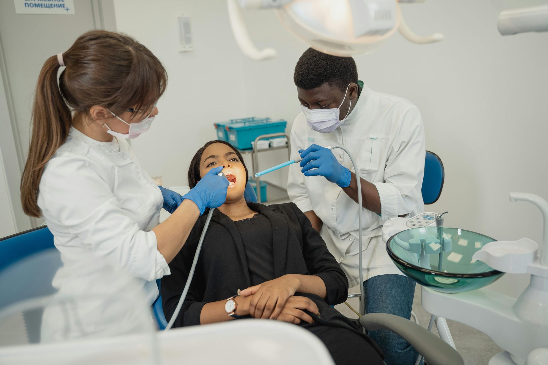 A patient receives a dental examination from a dentist and an assistant wearing scrubs, gloves, and masks in a clinic.