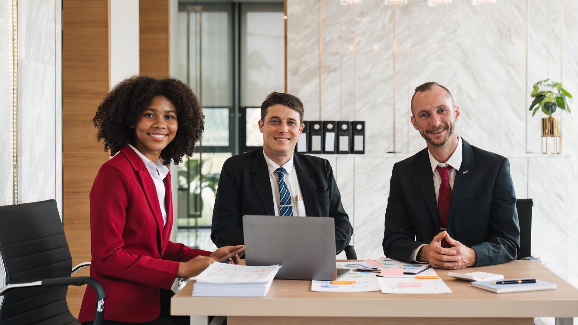 Three colleagues sit at a wooden desk with a laptop and documents in a bright, modern office, smiling at the camera.