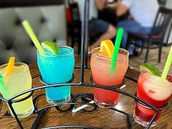 Four colorful cocktails with citrus garnishes in a metal drink holder, restaurant setting.