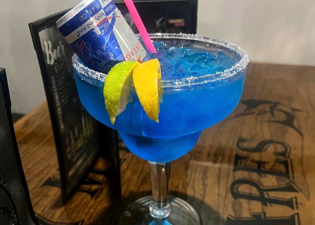 Blue cocktail in a margarita glass with a Red Bull can, lemon, lime, and a pink straw.