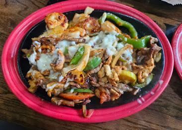 Sizzling fajitas with chicken, shrimp, peppers, and cheese on a black plate with a red rim.