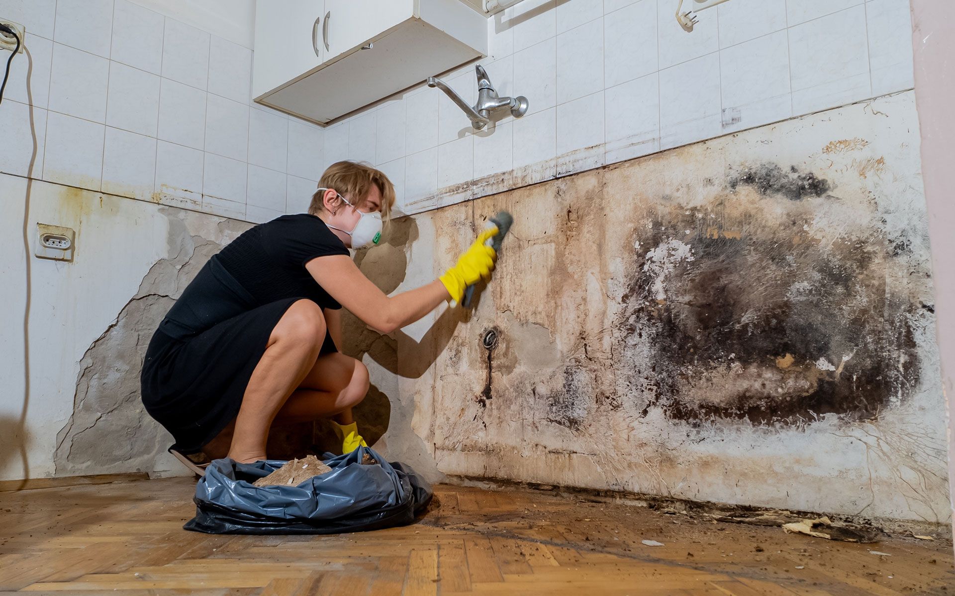 Person wearing a mask and gloves scrubbing a mold-infested kitchen wall.