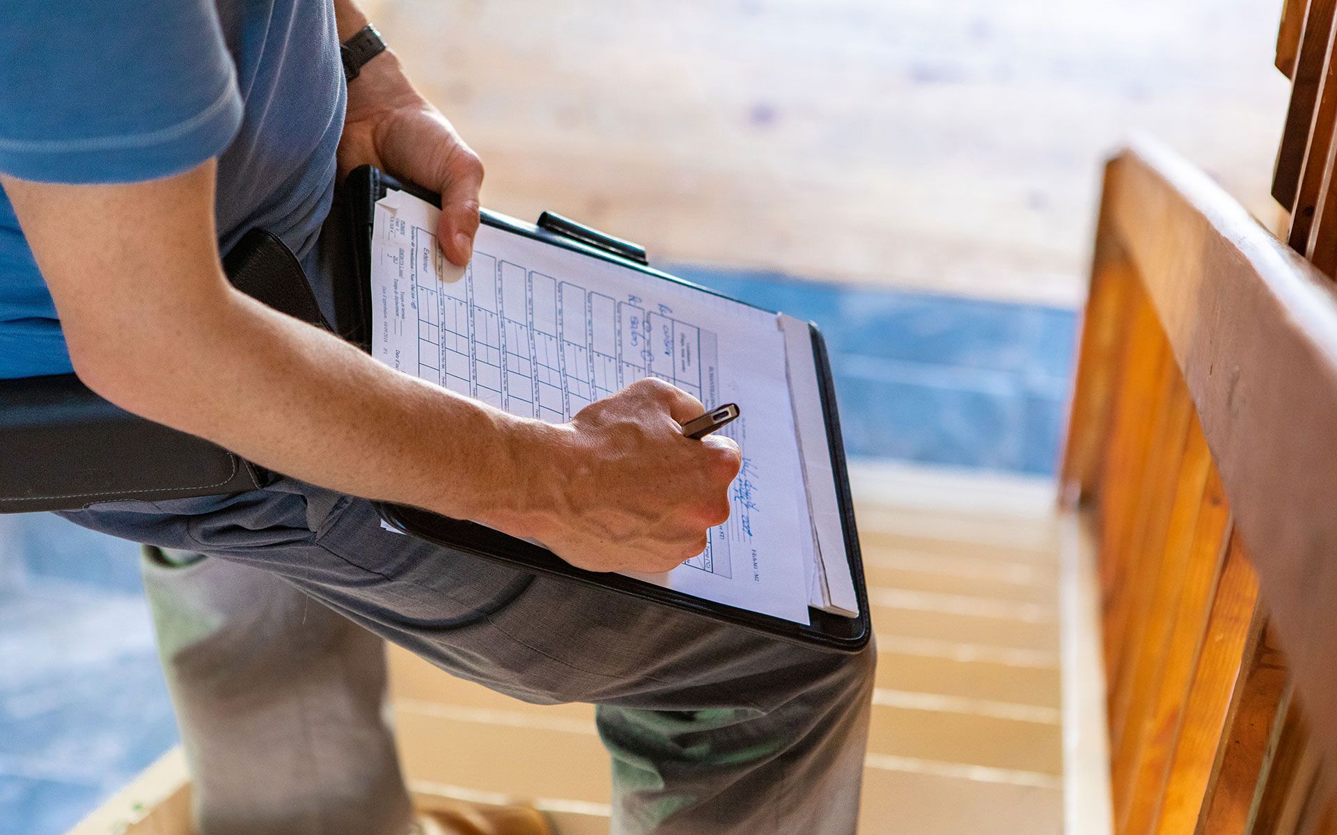 Person writing on clipboard, positioned on stairs. Blue shirt, jeans, wooden railing and steps are visible.
