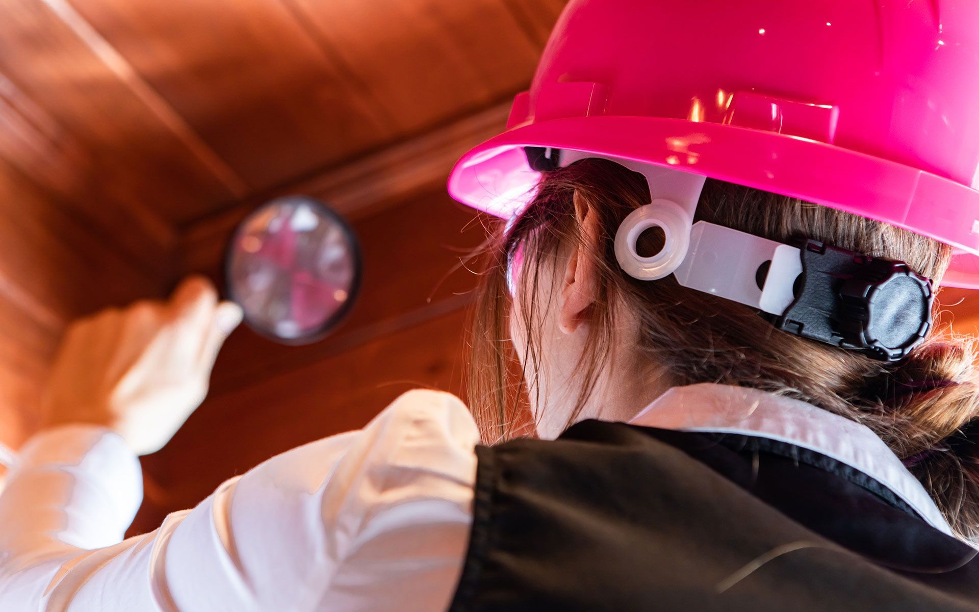 Person wearing a pink hard hat and headlamp, inspecting a surface with a magnifying glass.
