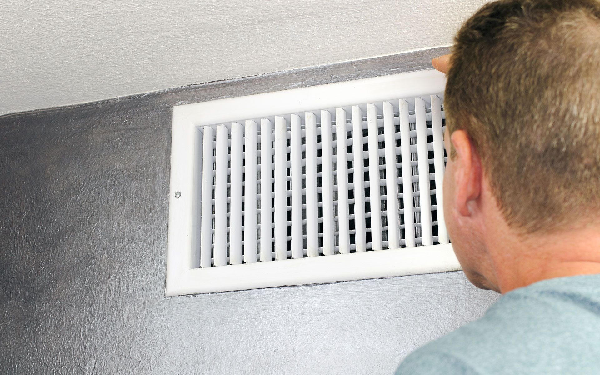 Man looking at a white air vent on a gray wall.