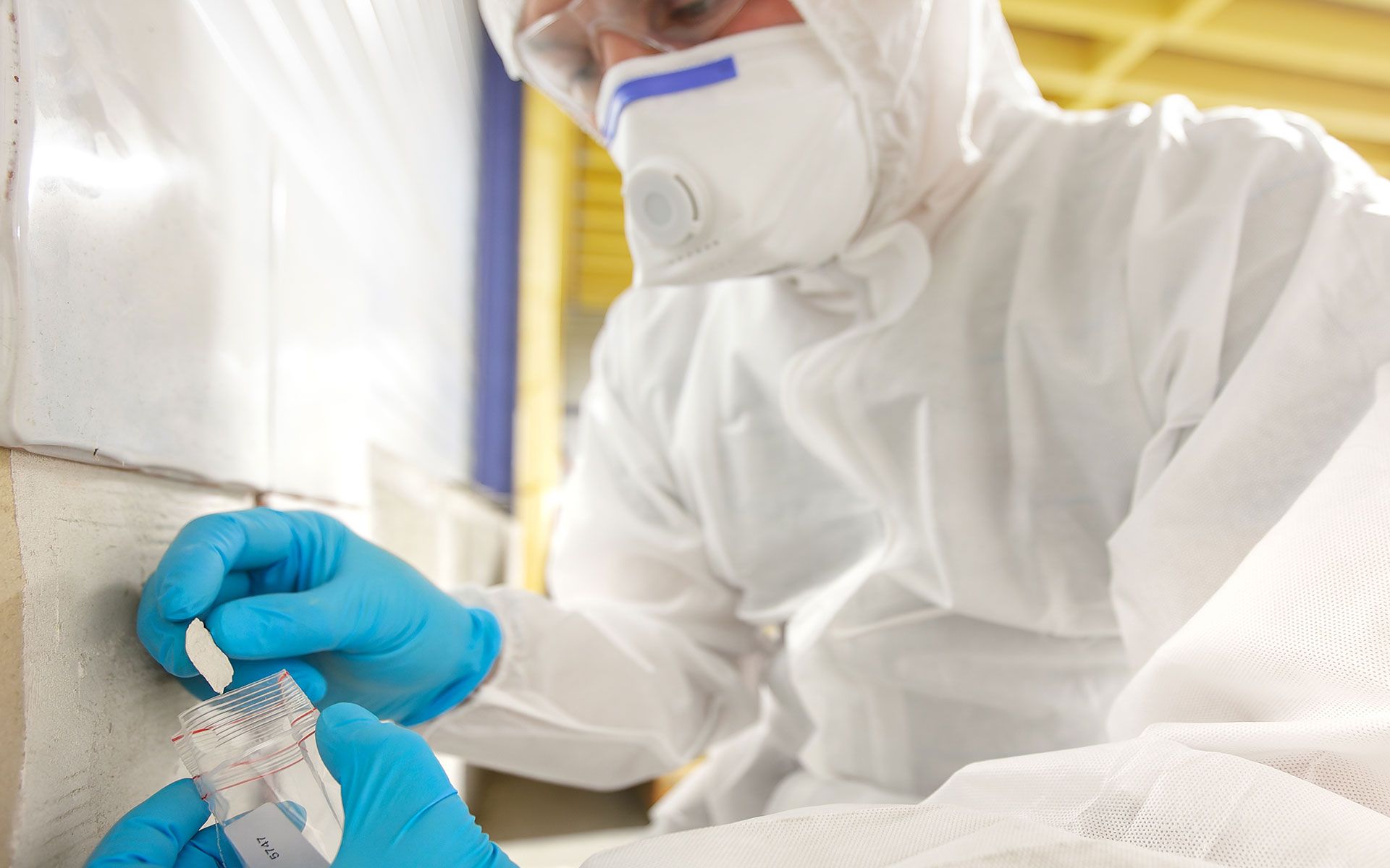 Person in protective suit taking a sample from wall, wearing mask and gloves.