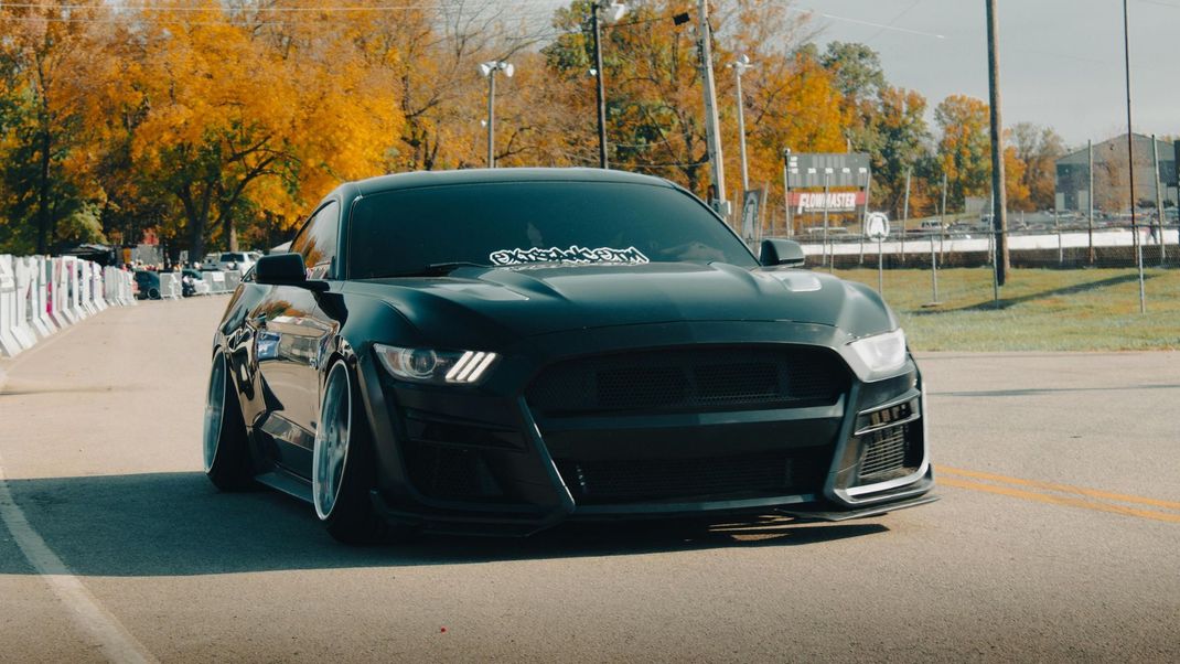 A black, lowered Ford Mustang with aftermarket wheels driving on an asphalt road during autumn.
