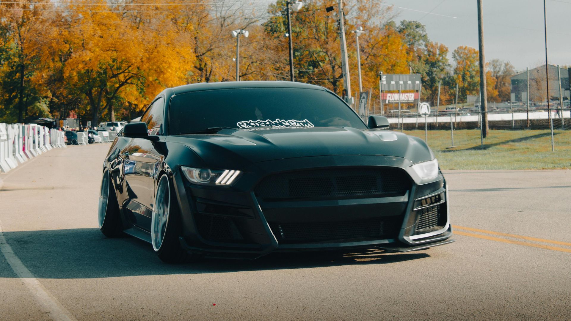 A black, lowered Ford Mustang with aftermarket wheels driving on an asphalt road during autumn.