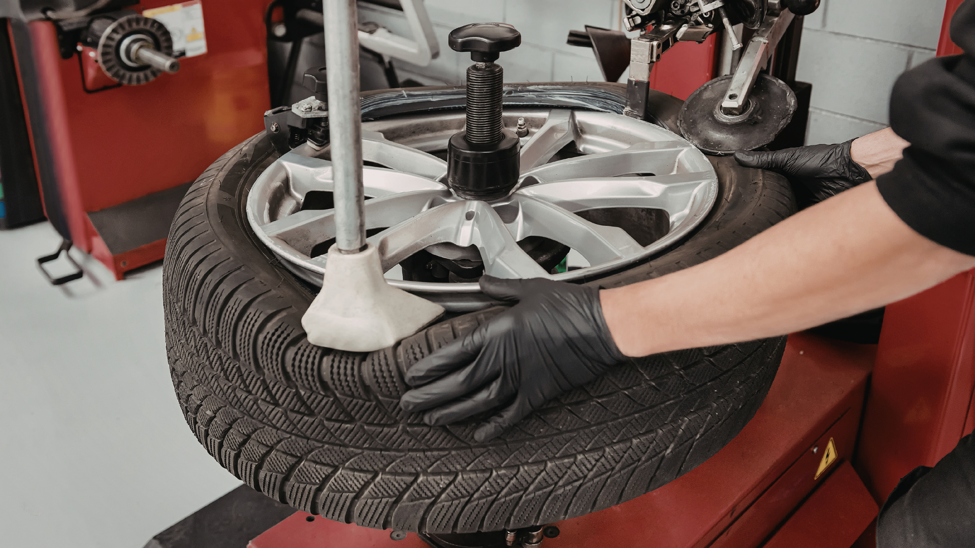 A technician in black gloves uses a tire changer machine to mount a tire onto a silver alloy car wheel.