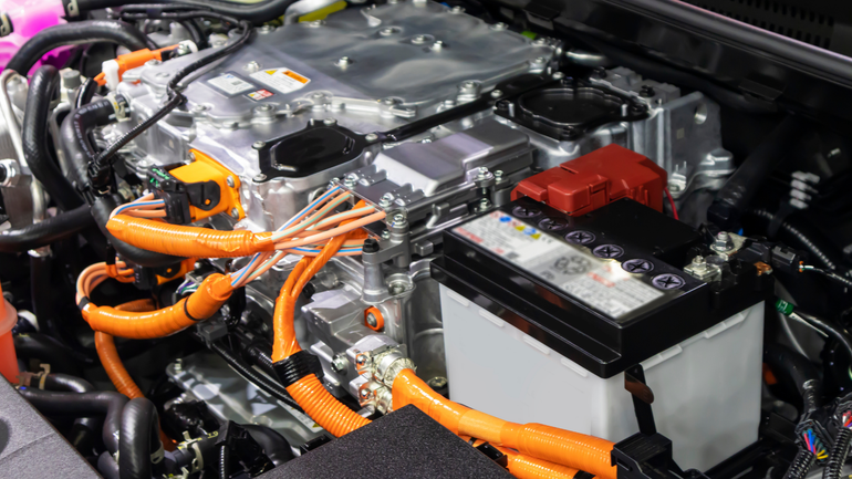 The engine bay of a hybrid vehicle featuring a 12-volt battery and high-voltage orange cables connected to an inverter.