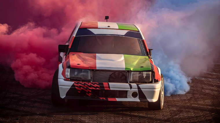 A car painted with the Irish flag colors drifts, surrounded by vibrant pink and blue smoke on a track.
