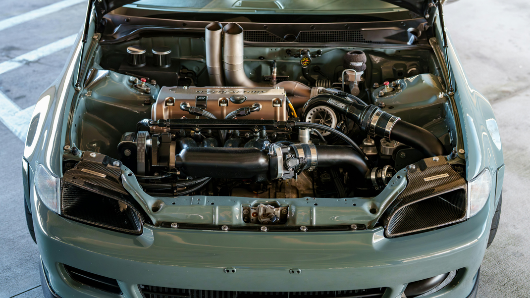 Open hood of a sage green Honda Civic showing a modified K-series engine with a prominent turbocharger and exhaust stack.