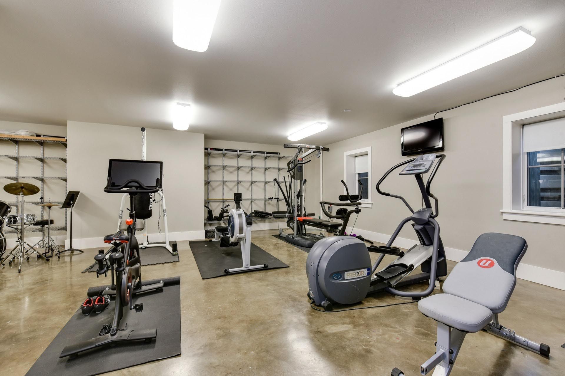 Home gym with various exercise machines, TVs, and equipment against neutral-colored walls.