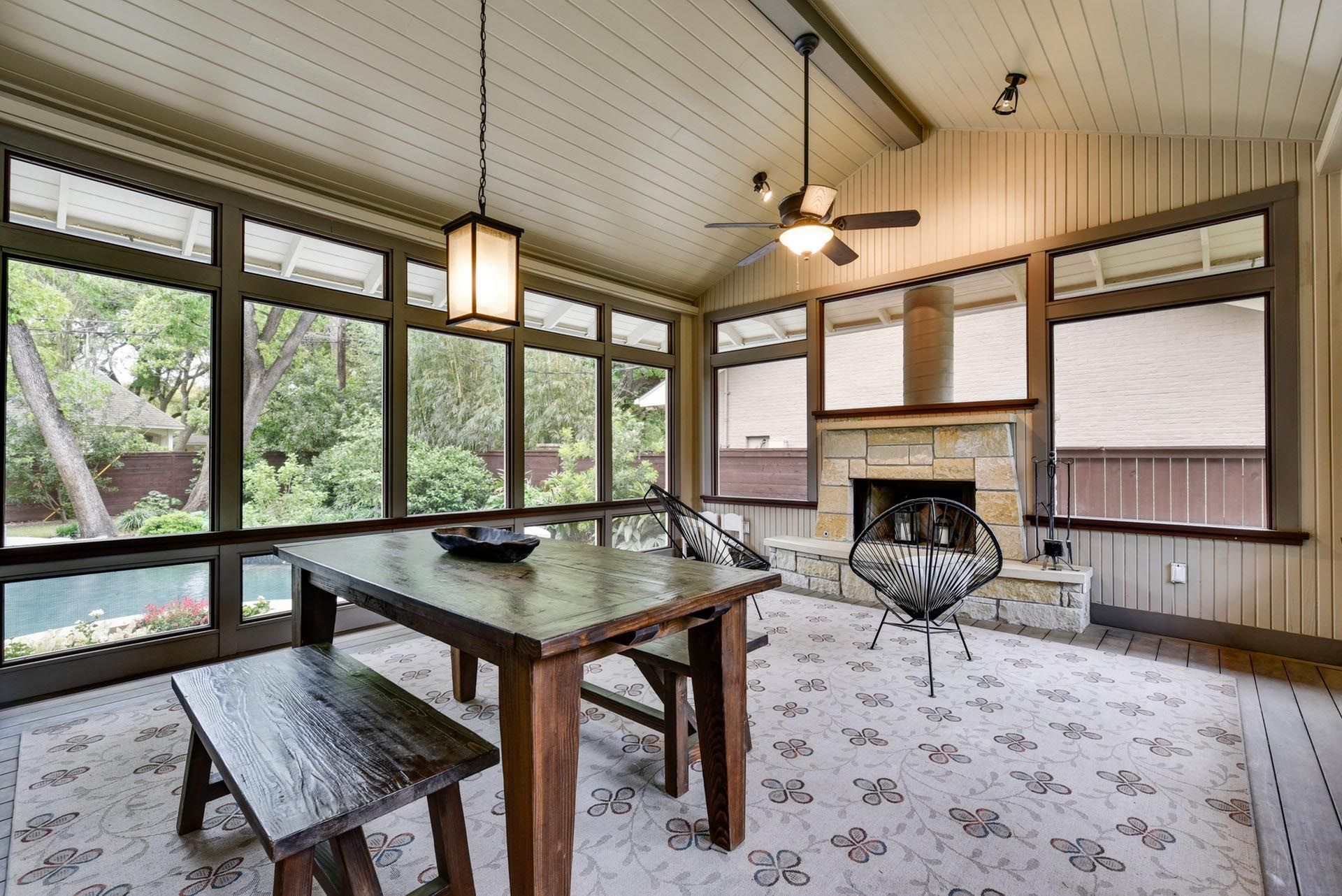 Sunroom with large windows, wooden table and bench, stone fireplace, and patterned rug.