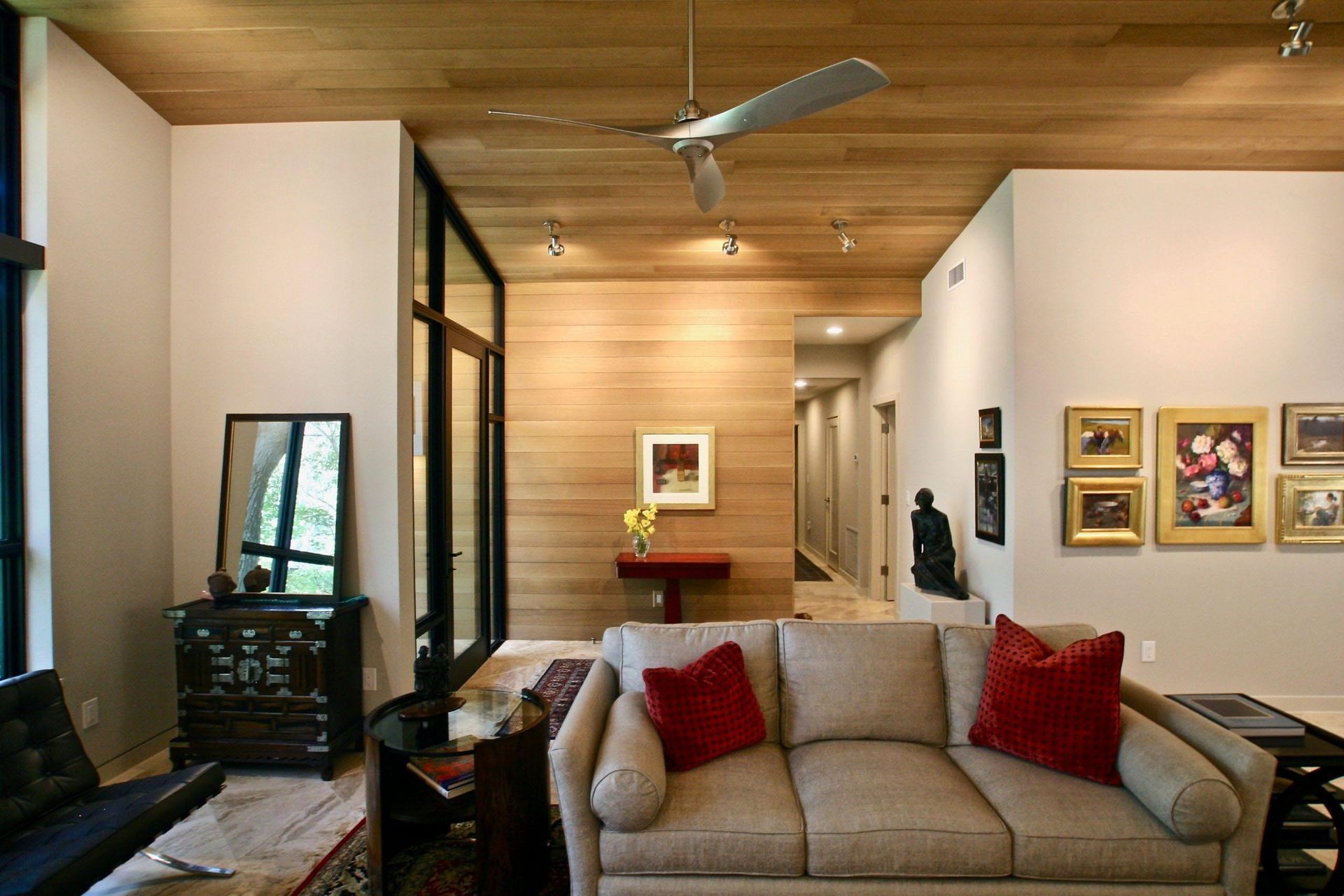 Living room with wood ceiling, tan walls, sofa, red pillows, art, and long hallway.