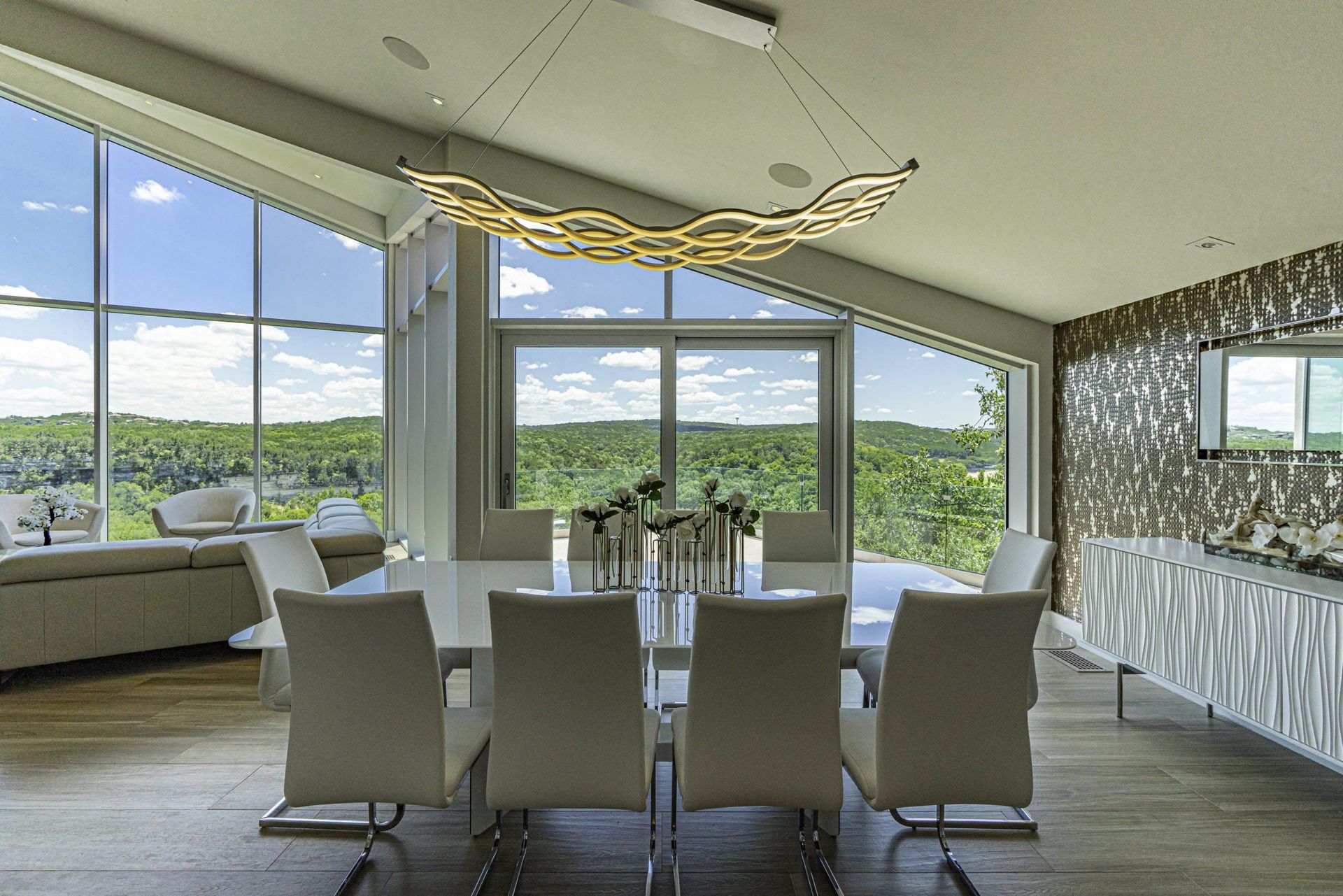 Modern dining room with a large window overlooking greenery. White table and chairs, abstract light fixture.