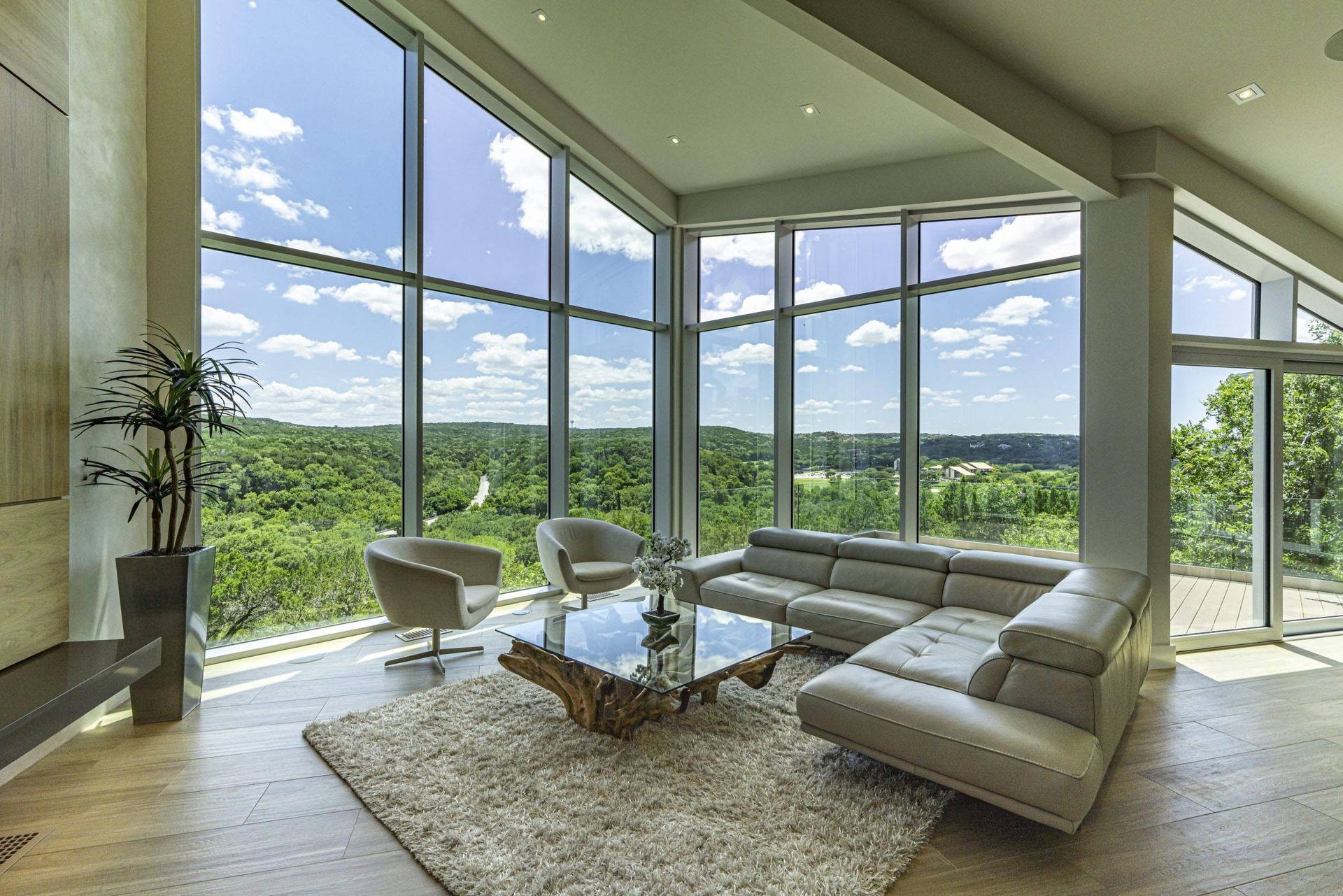 Modern living room with floor-to-ceiling windows overlooking a green landscape. Features a white sectional, armchairs