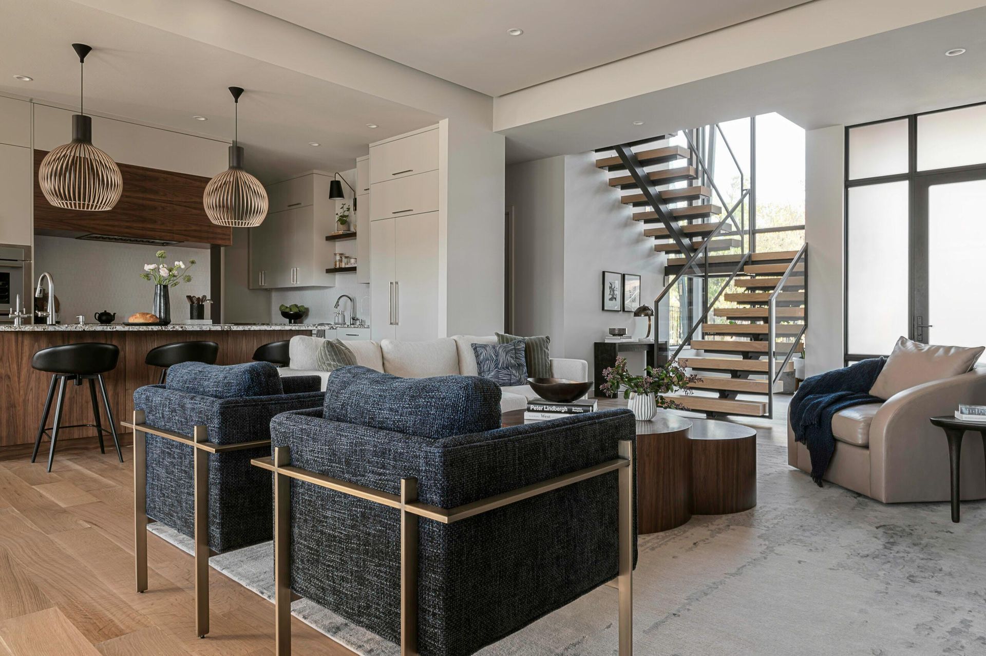 Modern living room with open kitchen, blue armchairs, and a floating staircase.