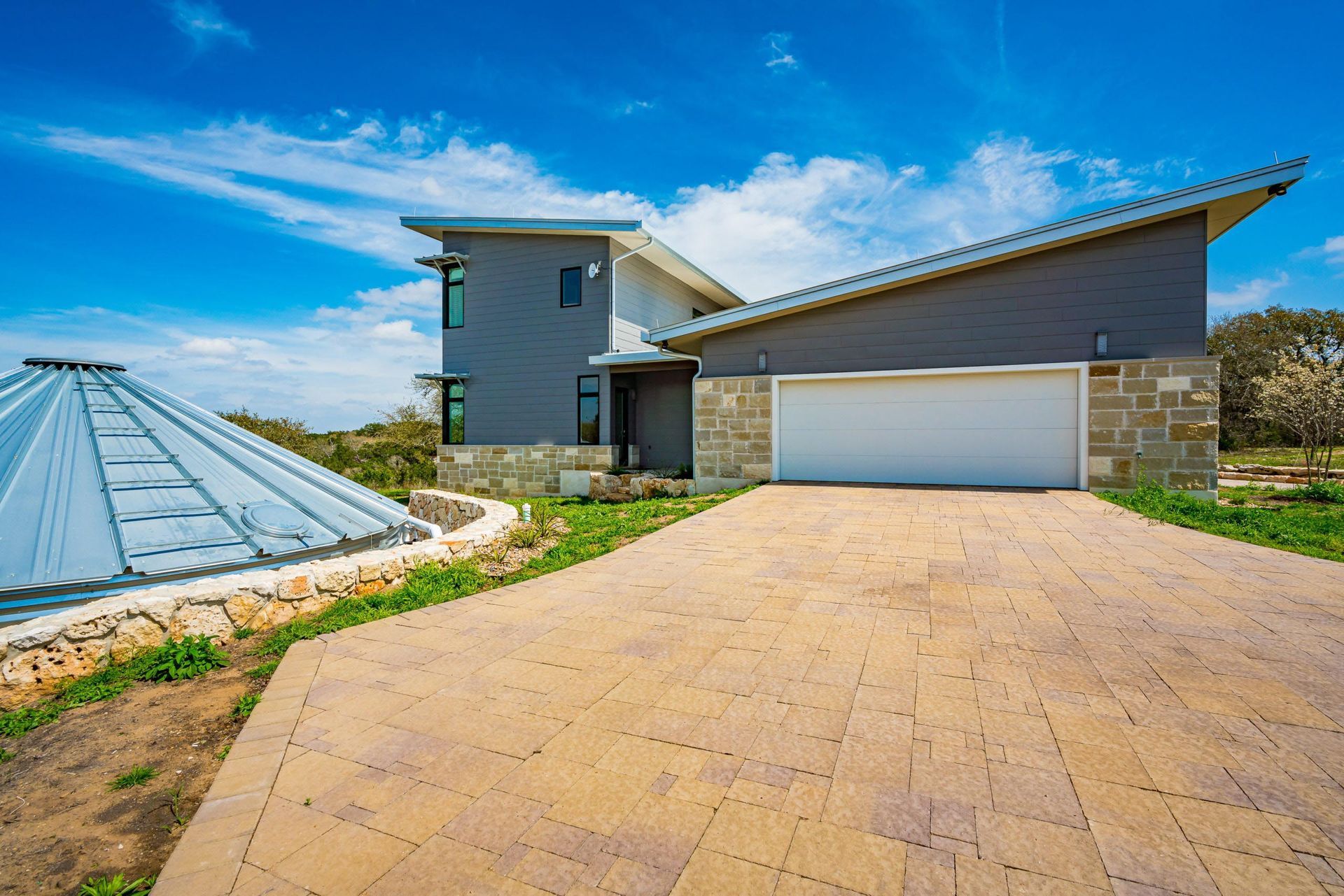 Modern gray house with a light-colored driveway and a blue sky. There is a garage and a dome-shaped structure.