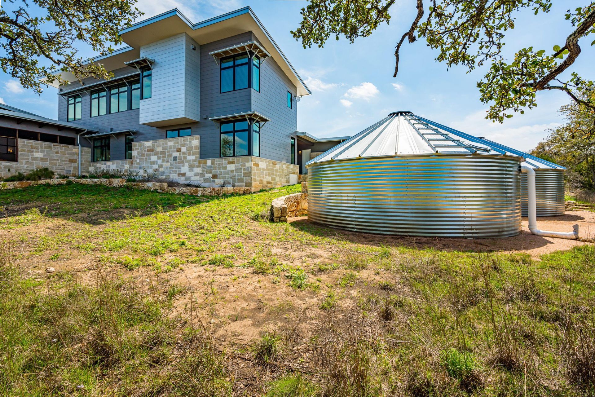 Modern home with blue and silver siding, next to galvanized metal water storage tanks on a grassy hillside.