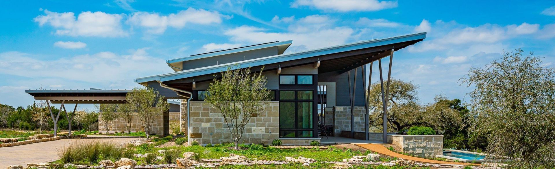 Modern house with a sloped roof under a blue sky, stone wall, and surrounding greenery.
