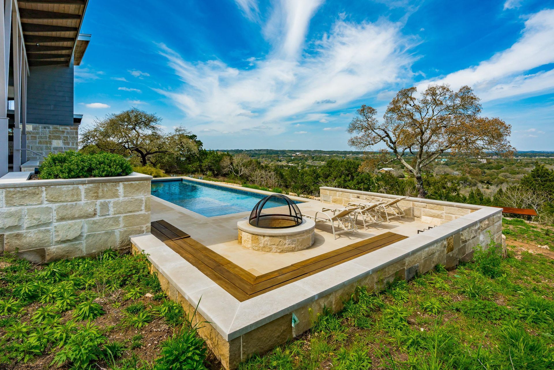 Pool area with fire pit, stone patio, and scenic view under a bright, partly cloudy sky.